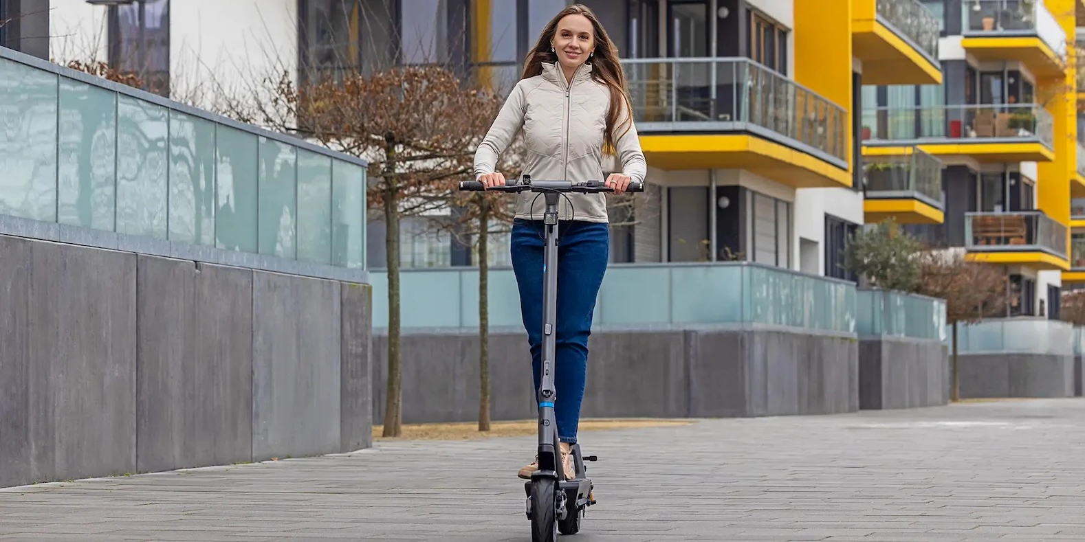s-Scooter kaufen Stadt Mobilitaet Frau fährt auf einem kompakten e-Scooter entlang moderner Wohngebäude und demonstriert nachhaltige Mobilität, Mikromobilität und Elektromobilität.