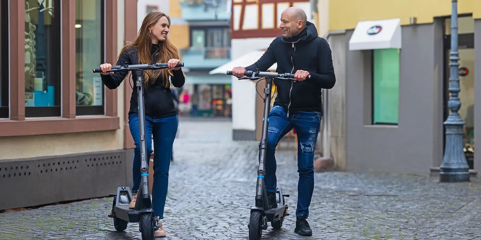 eScooter Mikro-Mobilitaet Zwei Erwachsene fahren nebeneinander auf Kopfsteinpflaster verschiedene Elektro-Scooter-Modelle. So testen Sie das Fahrkomfort der E-Scooter, um später eine Kaufentscheidung treffen zu können.