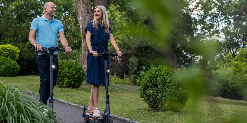 e-Scooter Reichweite Akku Zwei Erwachsene fahren nebeneinander auf E-Scootern im Radstreifen und demonstrieren sicheres Fahren mit Schutzhelm, angepasster Geschwindigkeit und vorausschauendem Bremsen im öffentlichen Straßenverkehr