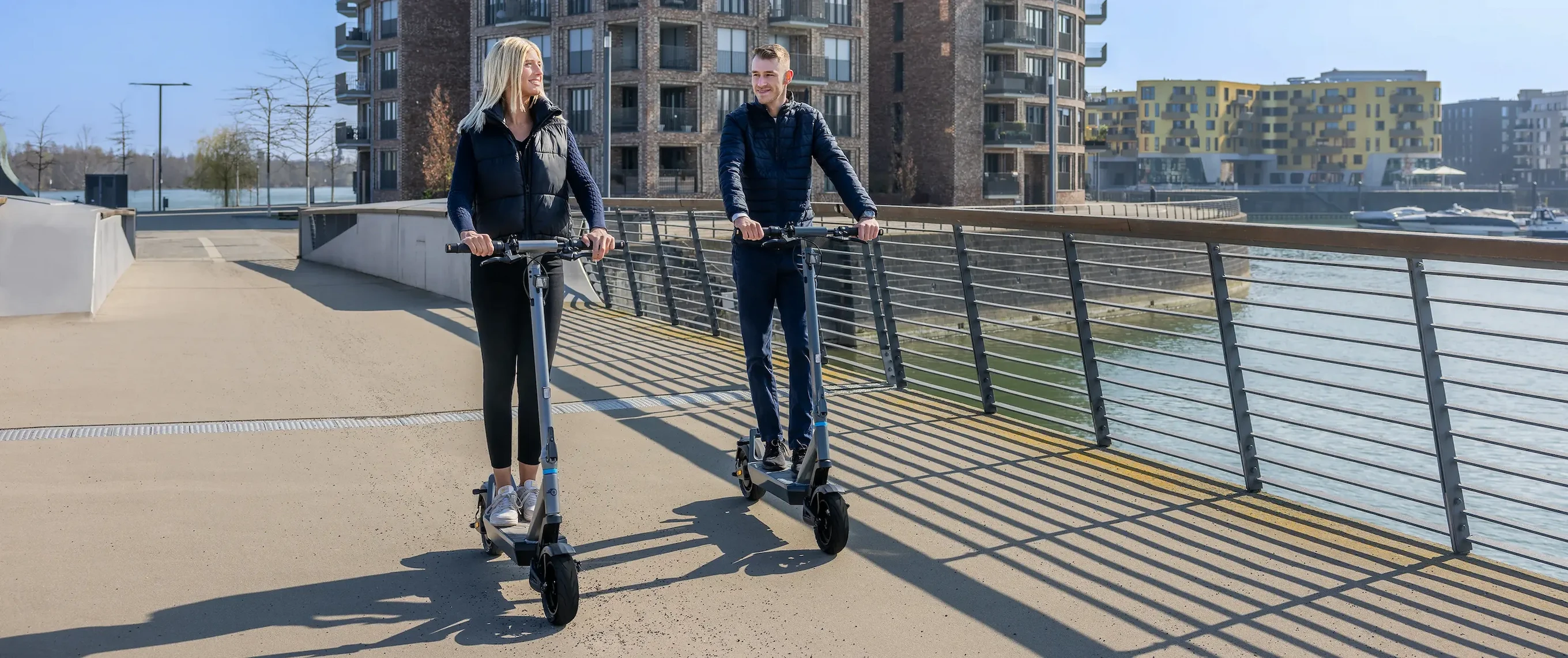 e-Scooter ePF Stadt Mobilitaet Zwei Erwachsene fahren auf kompakten e-Scootern entlang einer Uferpromenade und demonstrieren nachhaltige Mobilität, Mikromobilität und moderne Elektromobilität.